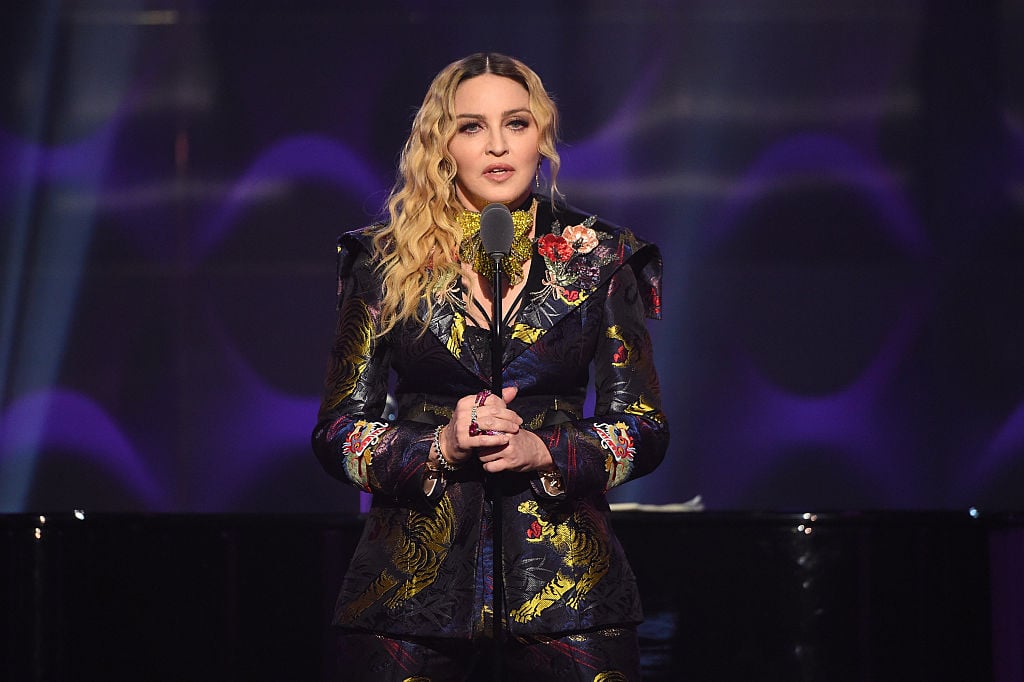 Madonna speaks on stage holding a microphone in front of a black and purple backdrop