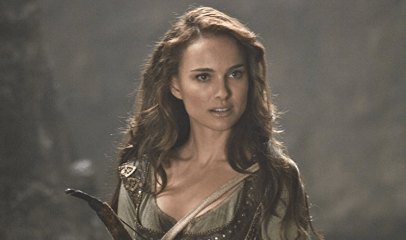 Natalie Portman holds a bow and arrow while standing in a forest. 