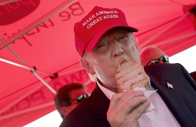 Donald Trump eats a pork chop on a stick wearing a red baseball cap.