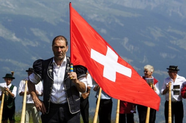 A flag thrower looks on as alphorn players prepare to perform in Nendaz, Switzerland. A flag thrower looks on as alphorn players prepare to perform in Nendaz, Switzerland.