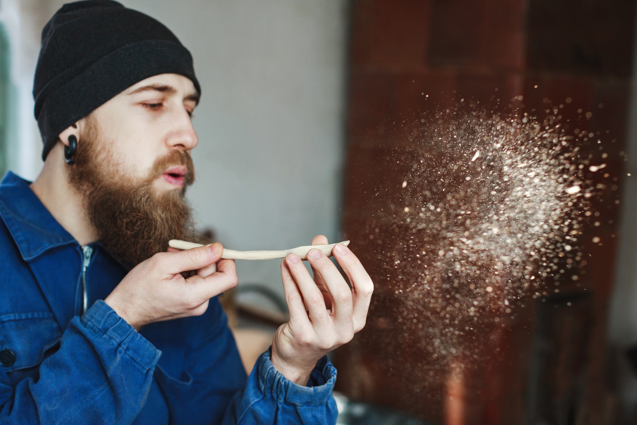 Man with a beard wearing blue jeans suit and black hat holding wooden spoon and blowing dust Man with a beard wearing blue jeans suit and black hat holding wooden spoon and blowing dust