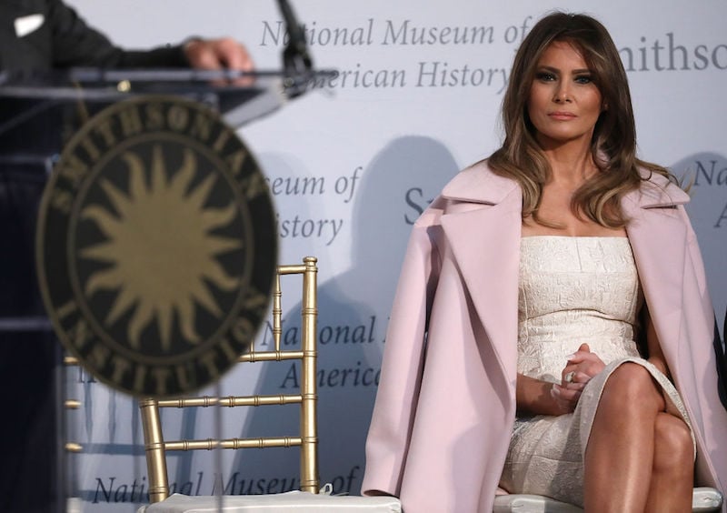 Melania Trump sitting while wearing a white dress and a pink coat. 