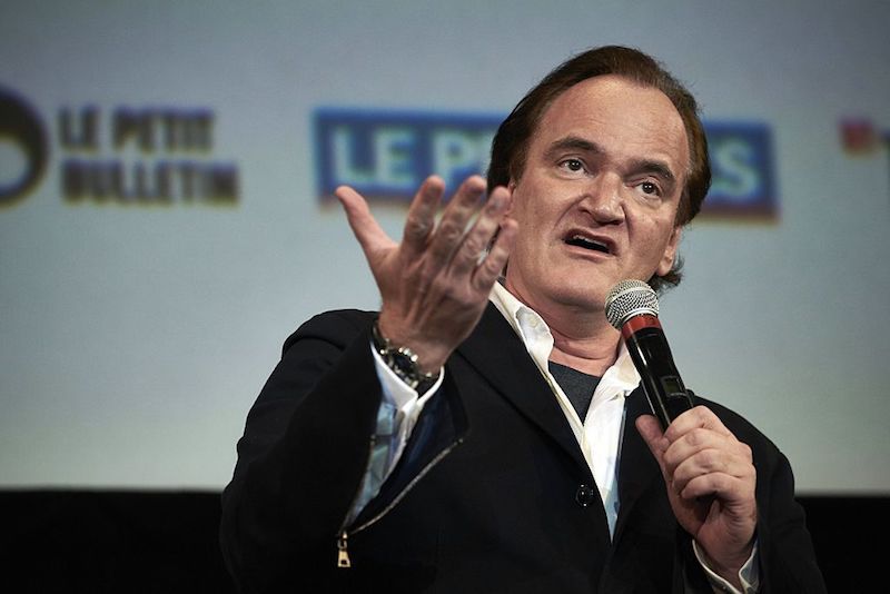 Quentin Tarantino holding a microphone and speaking on stage. Quentin Tarantino holding a microphone and speaking on stage.