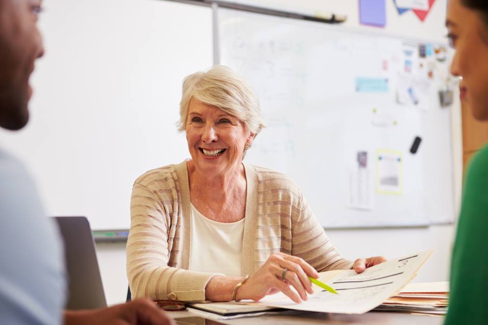 Senior teacher at desk talking to adult education students