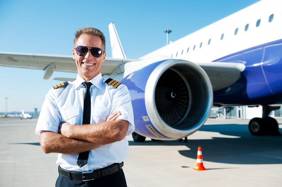 Confident male pilot in uniform keeping arms crossed and smiling with airplane in the background