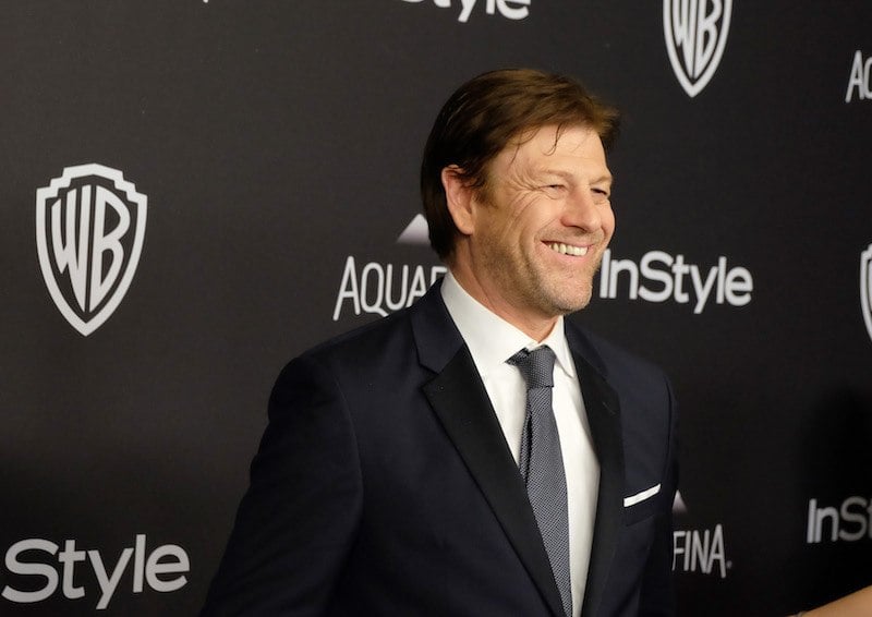 Sean Bean smiling while posing on a red carpet in a black suit and tie. 