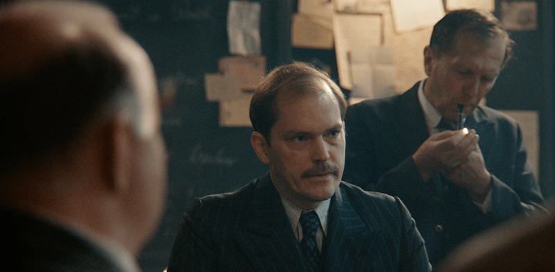 A man sits in a suit surrounded by other men. A man sits in a suit surrounded by other men.