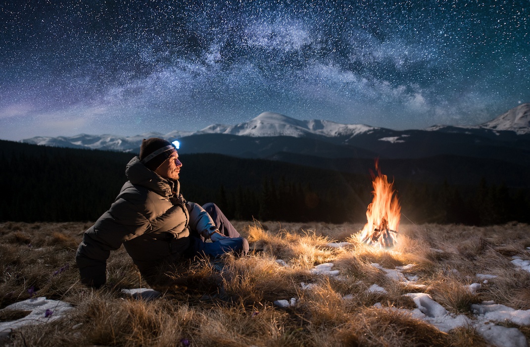 Male tourist have a rest in the mountains at night. Guy with a headlamp sitting near campfire under beautiful night sky full of stars and milky way, and enjoying night scene