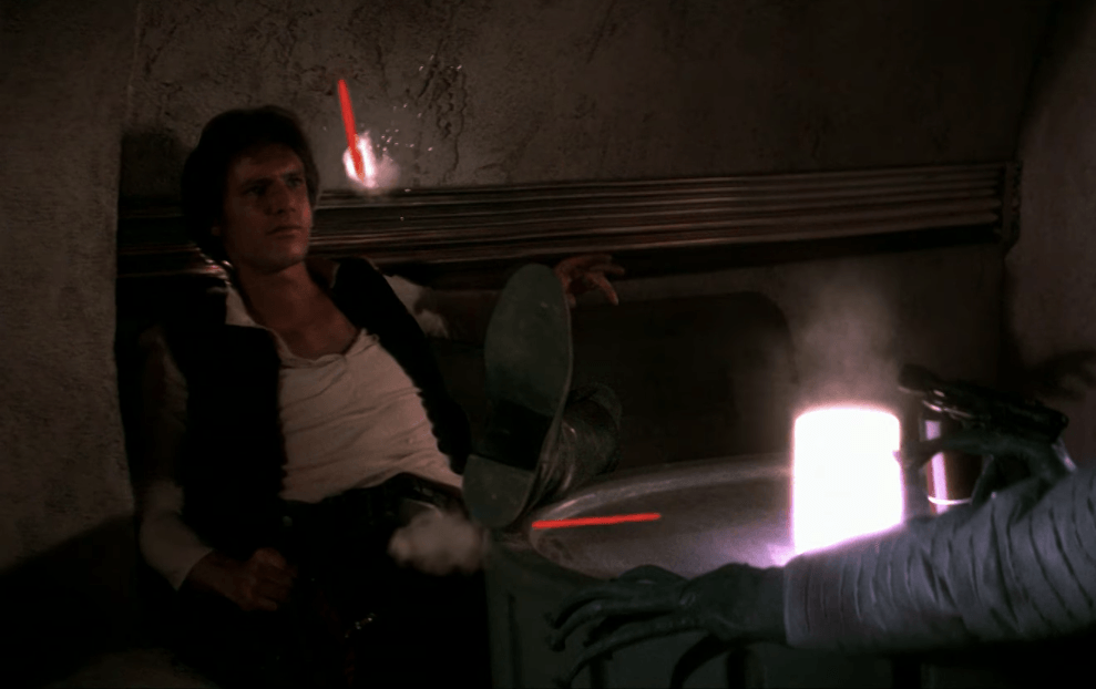 Greedo shoots Han as he sits in front of him. 