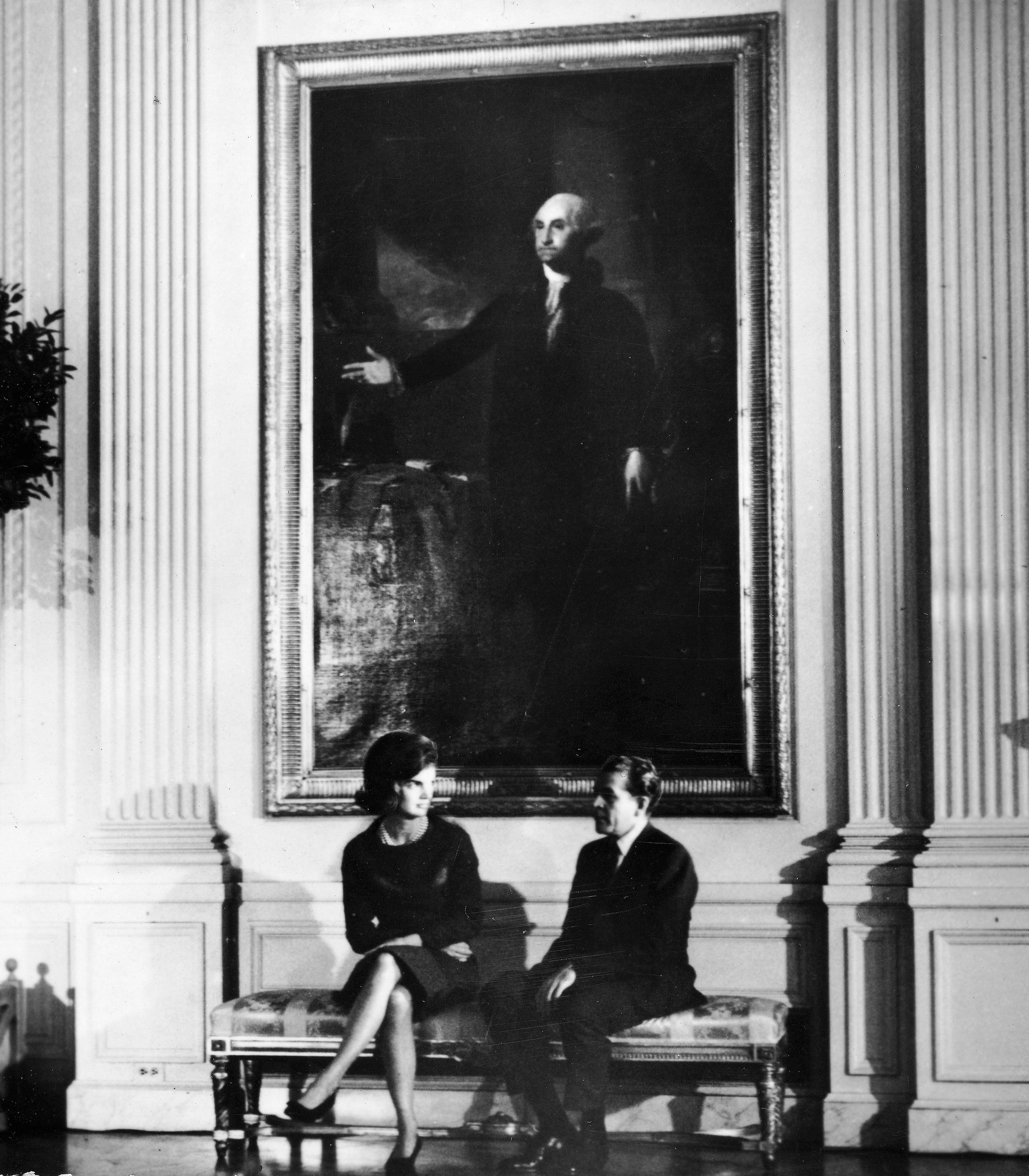 Jacqueline Kennedy and Charles Collingwood sit on a bench below the painting of President George Washington, during the televised tour of the White House