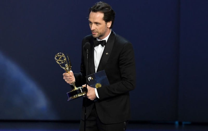 Matthew Rhys wearing a black tuxedo, holding an Emmy award, and speaking into a microphone.
