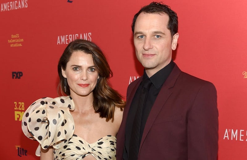 Keri Russel wearing a polka dot dresses stands next to Matthew Rhys, wearing a red jacket and black shirt.