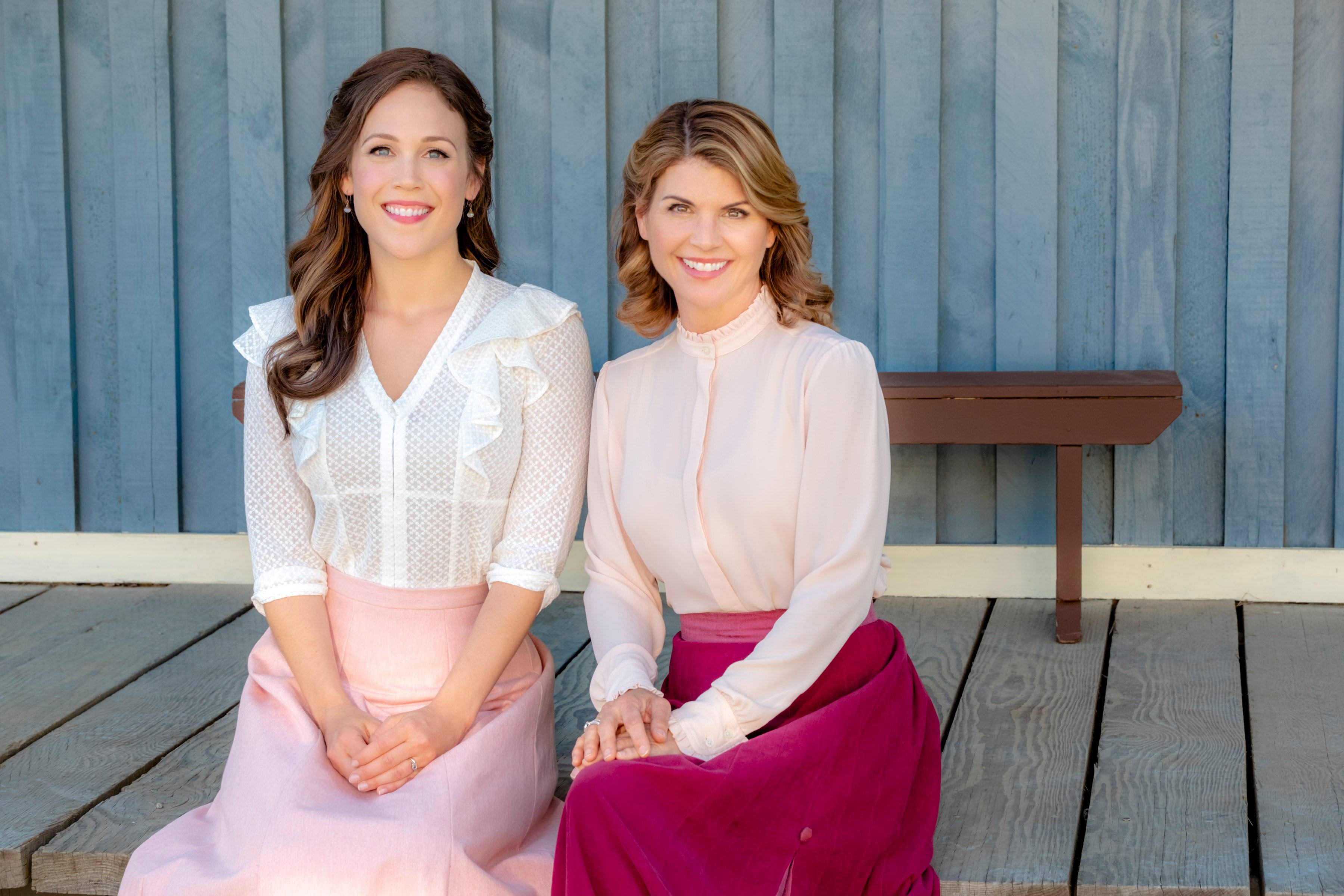 Erin Krakow and Lori Loughlin sitting next to each other on a wooden sidewalk in 'When Calls the Heart' 