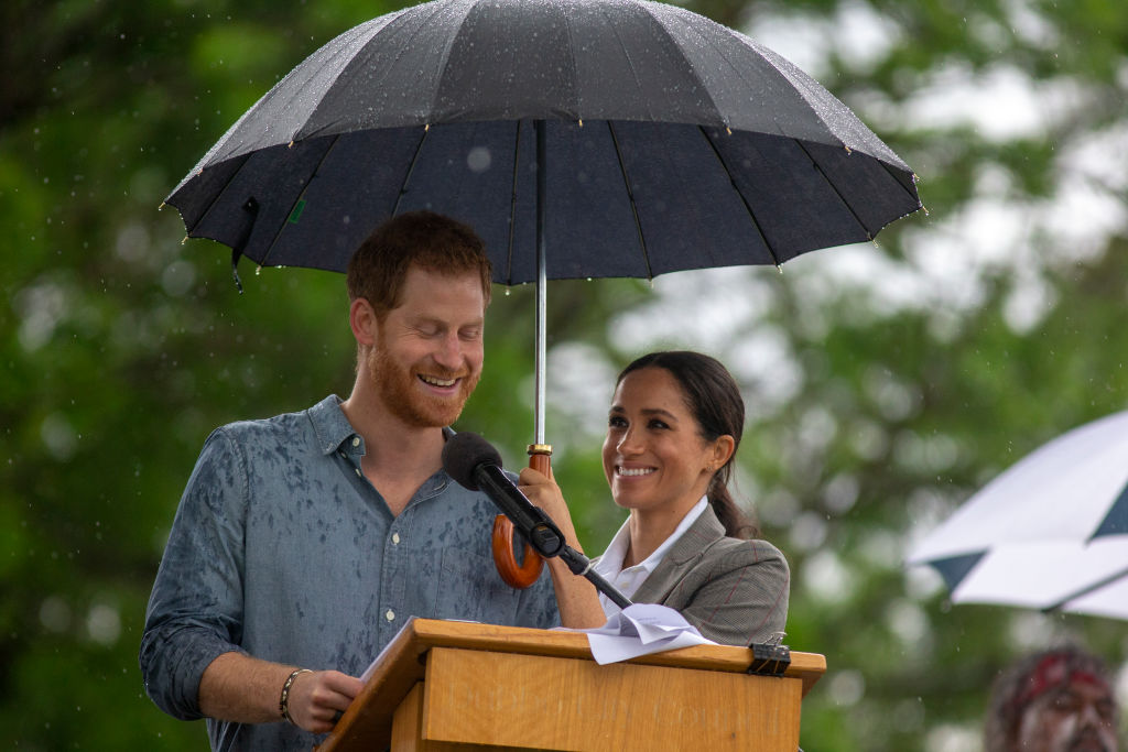 Prince Harry gives speech while Meghan Markle holds umbrella.