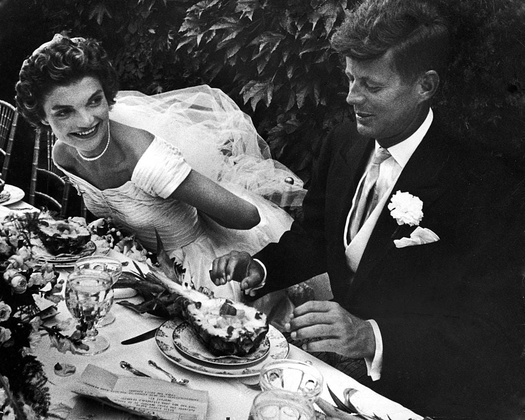 Senator John Kennedy & his bride Jacqueline in their wedding attire, as they sit down together at table to begin eating a pineapple salad at formally set table outdoors at their wedding reception.