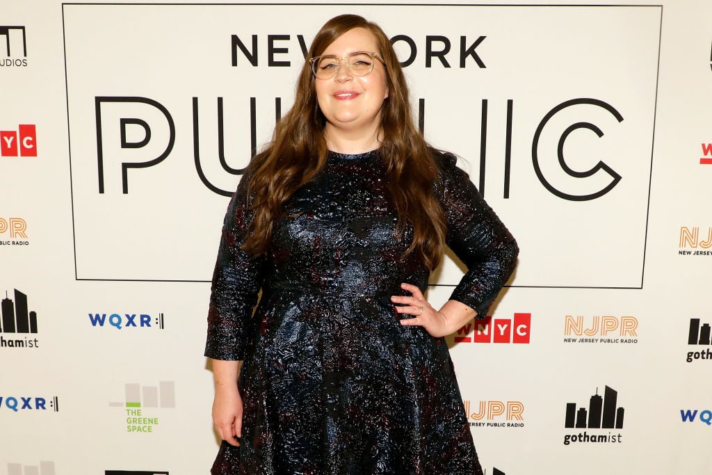 Aidy Bryant in a dark blue dress smiling in front of a repeating background.