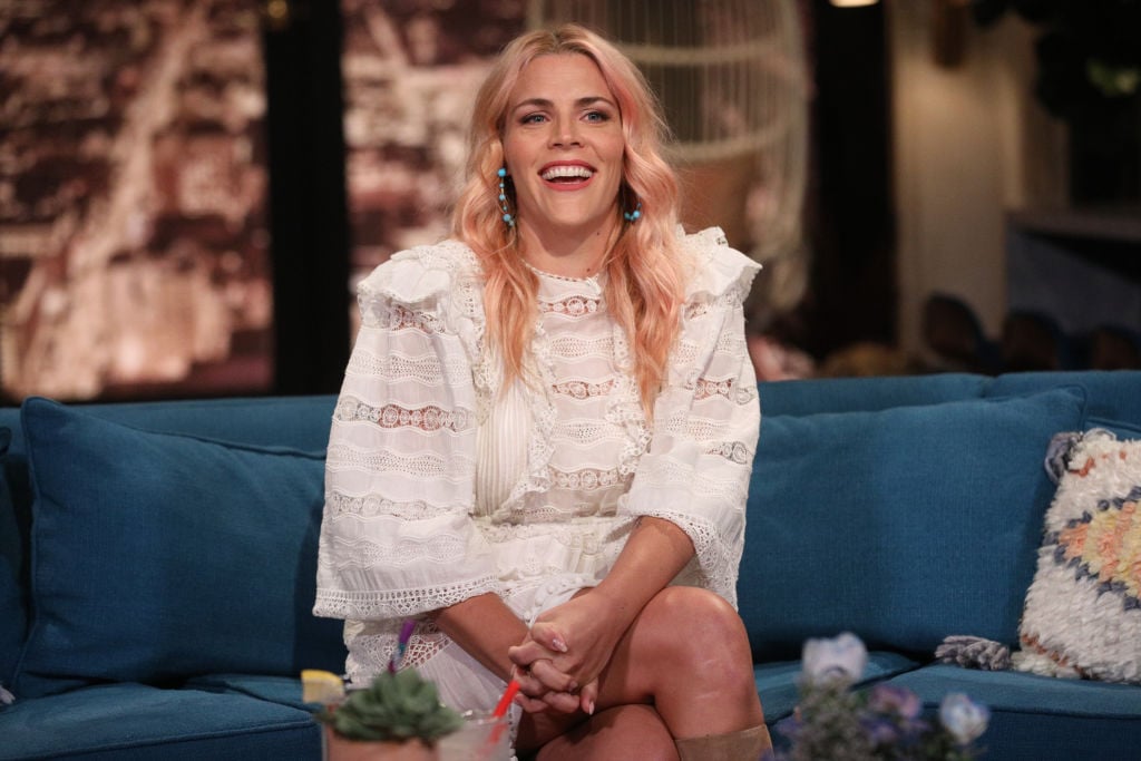 Busy Philipps in a white dress sitting on a couch laughing