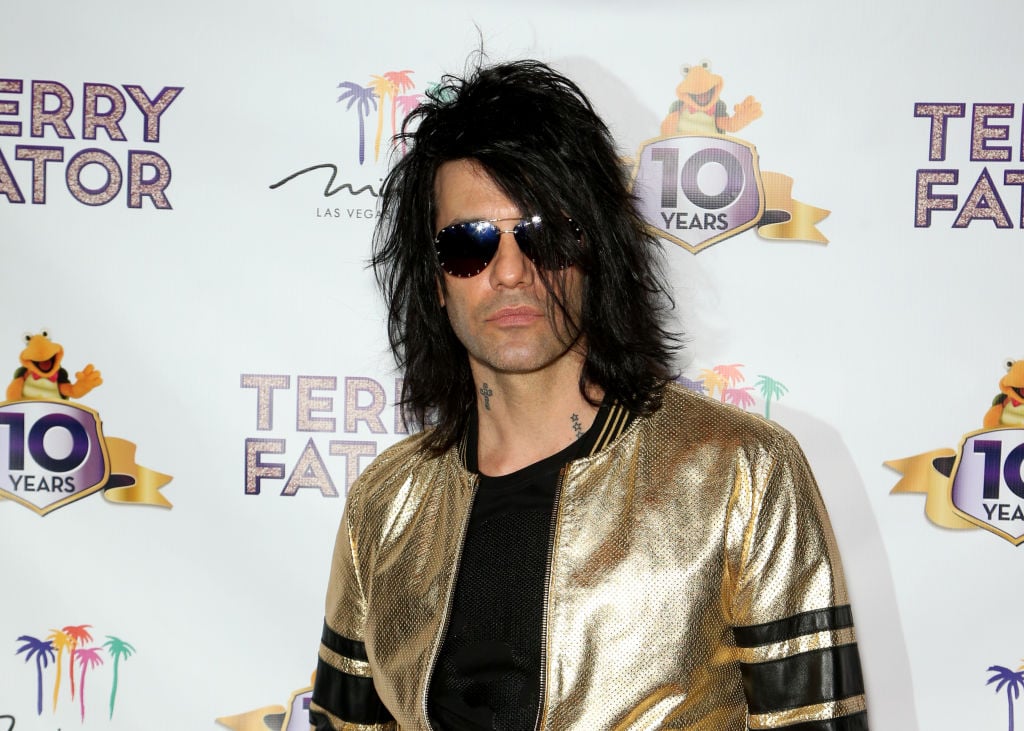 Criss Angel in sunglasses and a gold jacket in front of a repeating background