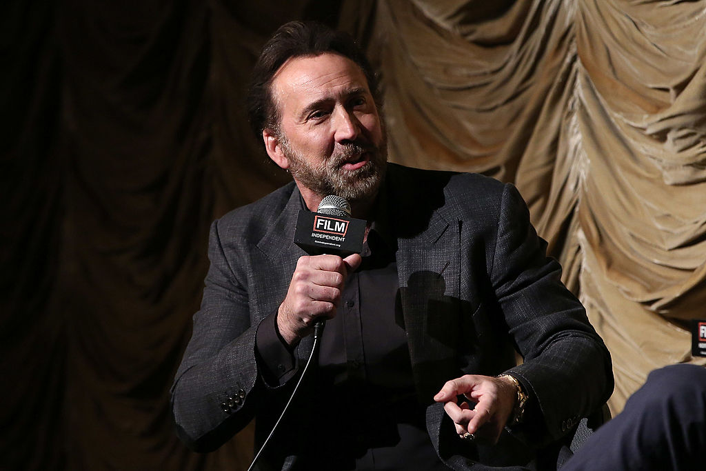 Nicolas Cage holding a microphone in front of a velvet yellow curtain