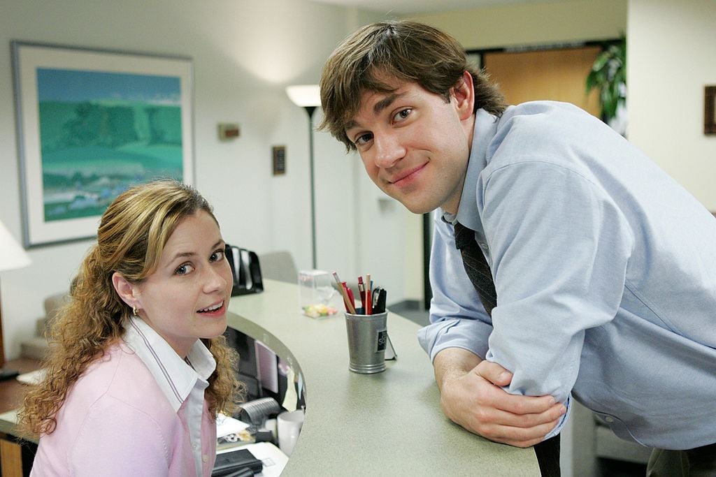 Pam Beesly (Jenna Fischer) of 'The Office' sits behind her reception desk while Jim Halpert (John Krasinski) leans over it.