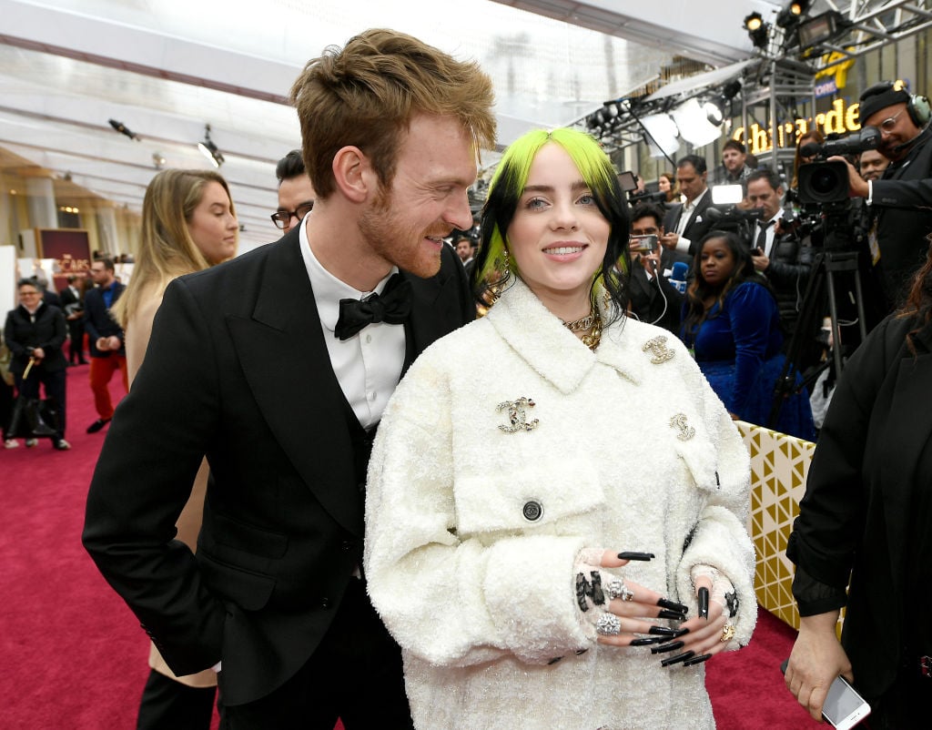 Billie Eilish wearing a white collared Chanel jacket standing in front and to the right of her brother, Finneas O'Connell