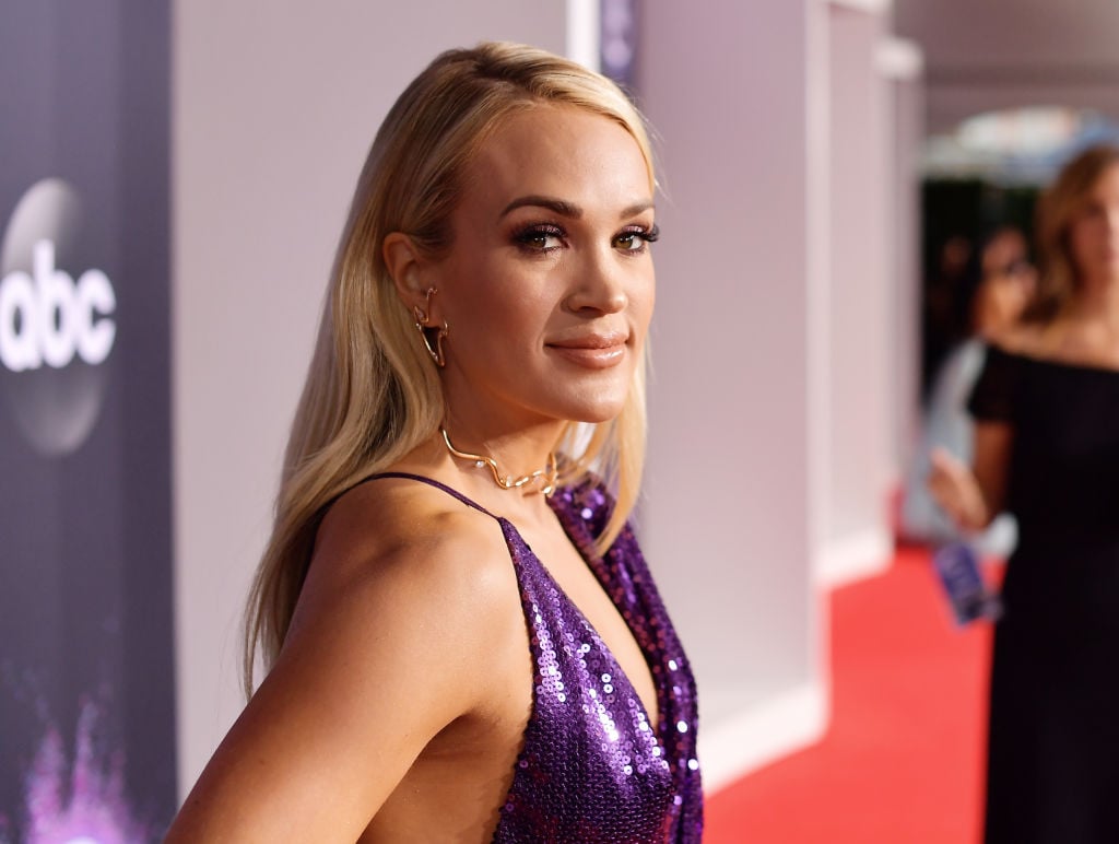 Carrie Underwood in purple, smiling at the camera in front of a repeating background