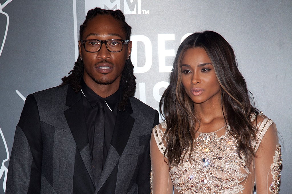 Future (left), wearing glasses and a black suit with Ciara (right) looking off camera in a gold jeweled dress