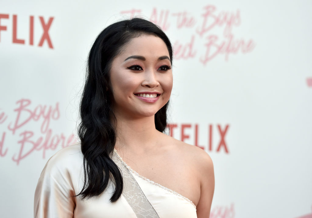 Lana Condor wearing a white one shoulder dress, standing in front of a repeating background