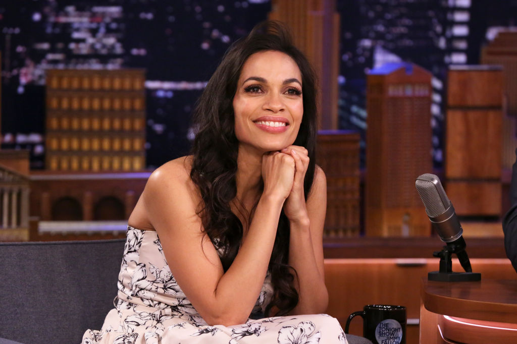 Rosario Dawson, sitting and smiling with her face resting on her hands