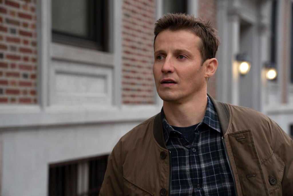 Will Estes looking off camera in a blue flannel shirt and brown jacket