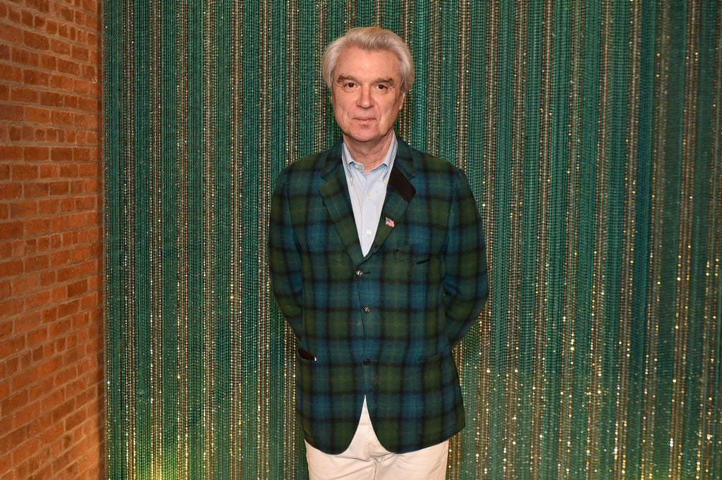 David Byrne smiling in front of a blue-green backdrop