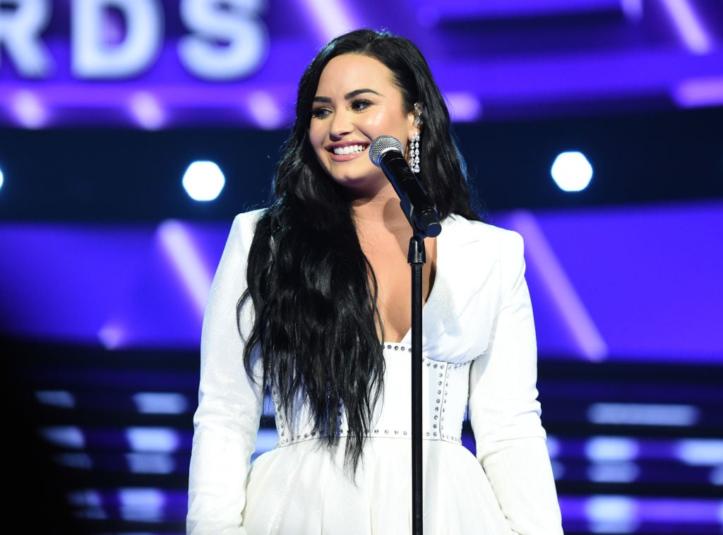 Demi Lovato smiling in a white dress, standing on a stage in front of a microphone