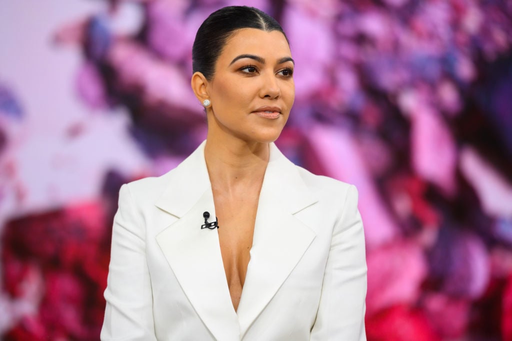 Kourtney Kardashian in a white blazer in front of a pink and purple background