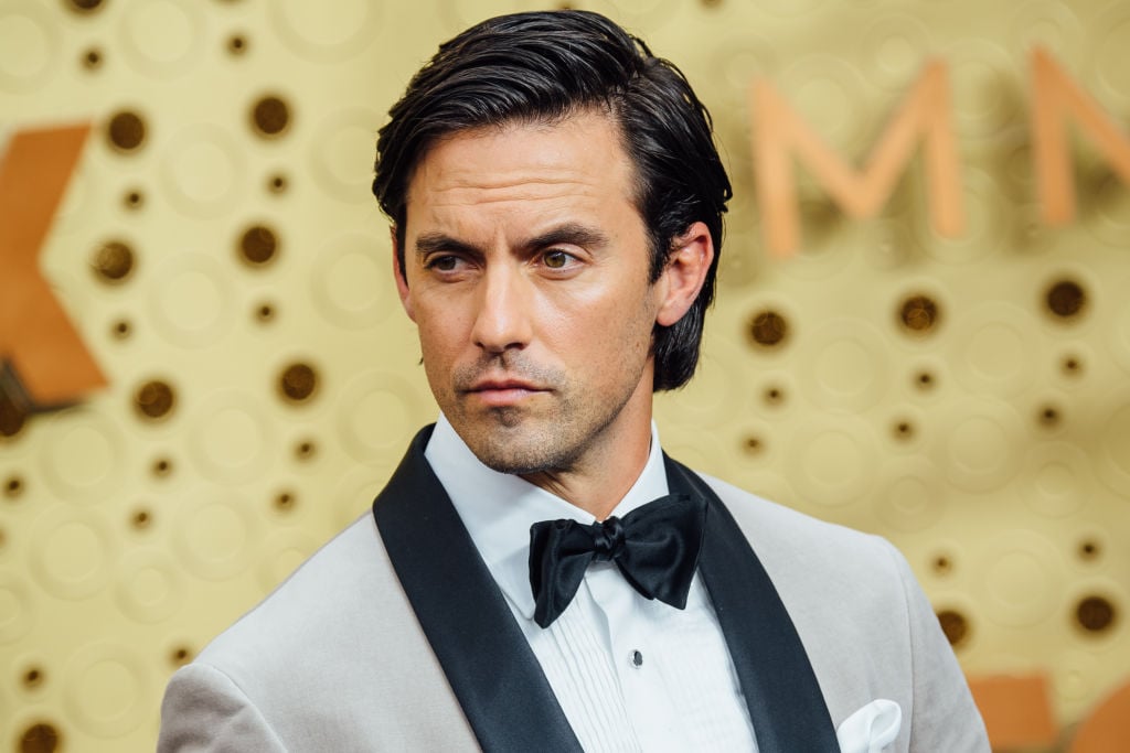 Milo Ventimiglia standing in front of a repeating background in a white and black jacket and black bowtie