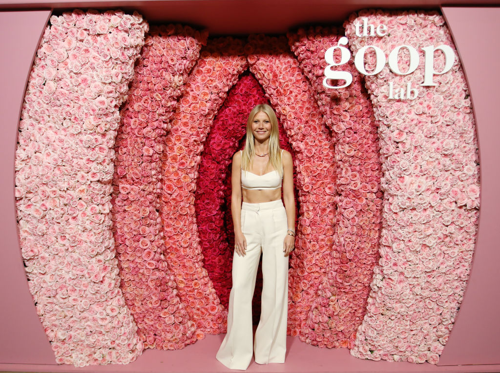 Gwyneth Paltrow posing in front of a flower wall that says "the goop lab"