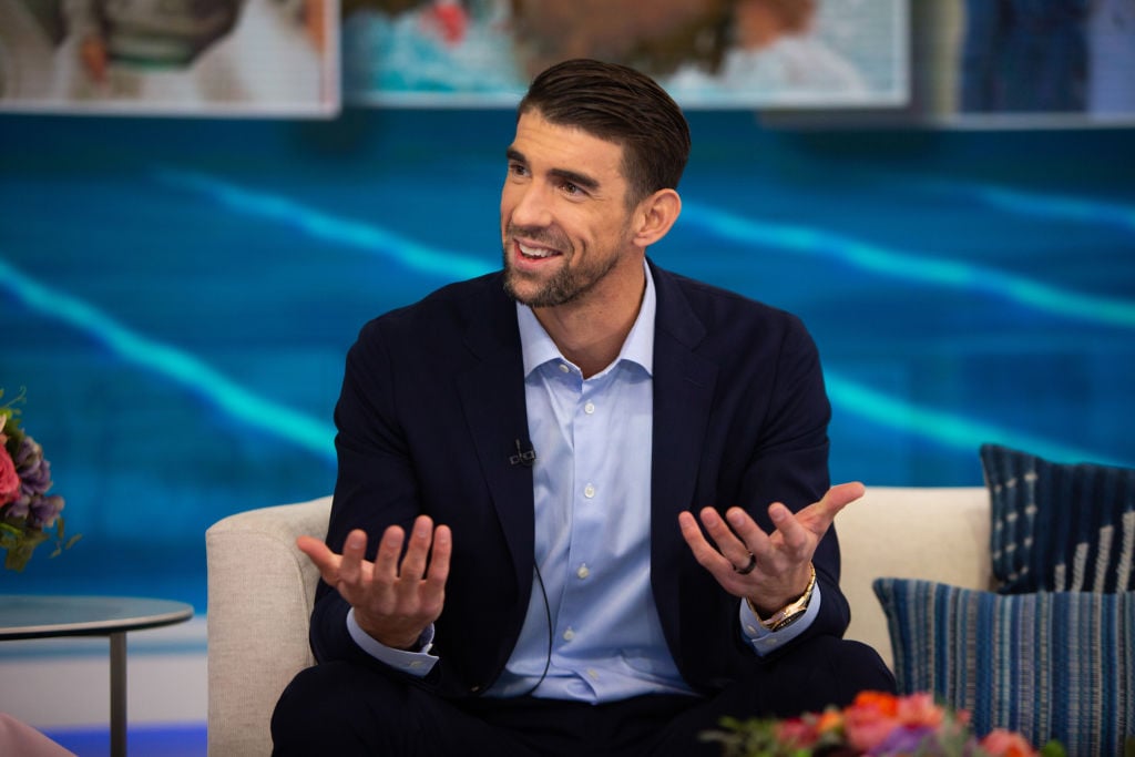 Michael Phelps, seated, smiling in front of a blue background