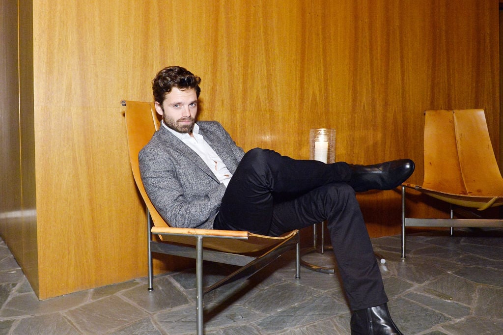 Sebastian Stan smiling, reclined in a chair in front of a wood paneled background