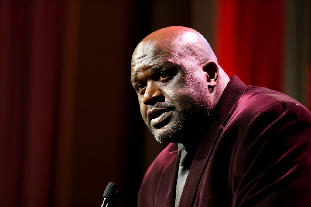 Shaquille O'Neal at a podium in a dark red jacket