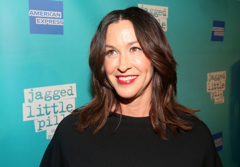 Alanis Morissette smiling turned slight away from the camera in front of a blue repeating background