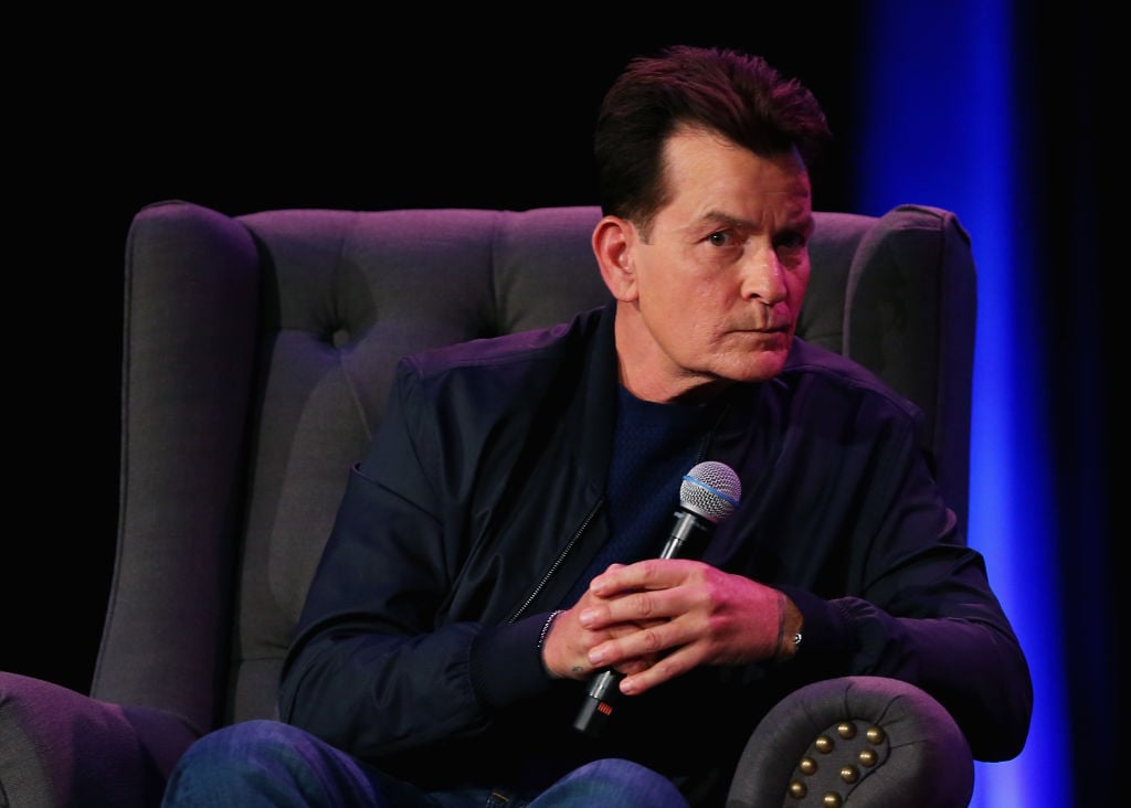 Charlie Sheen sitting in a chair holding a microphone looking away from the camera