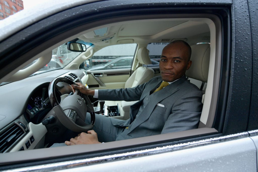 A chauffeur in a gray suit driving a Lexus, looking out the window at the camera