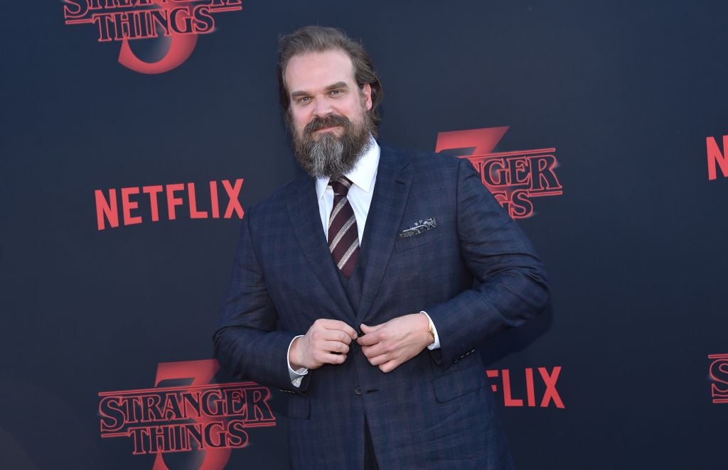 David Harbour buttoning his jacket, smiling, in front of a repeating background with the 'Stranger Things' logo