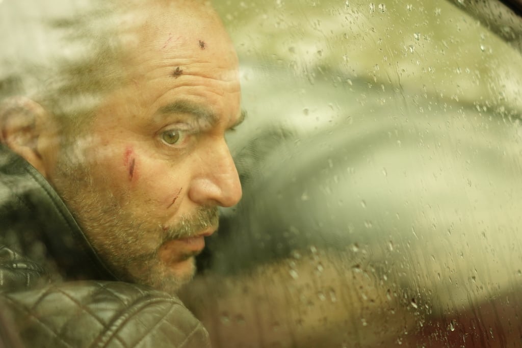 Production still with a sepia tone and a man with cuts on his face looking through a car window with rain on the glass
