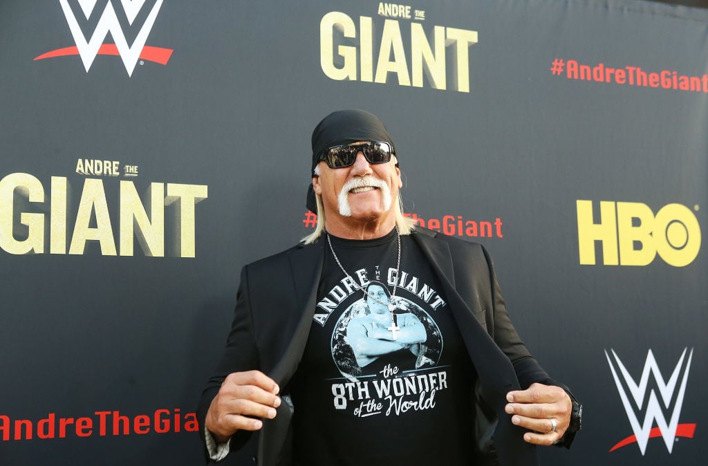 Hulk Hogan smiling in front of a black background with a repeating logo