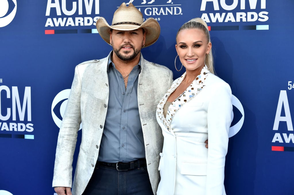 Jason and Brittany Aldean smiling at the camera in front of a blue backdrop with repeating logos