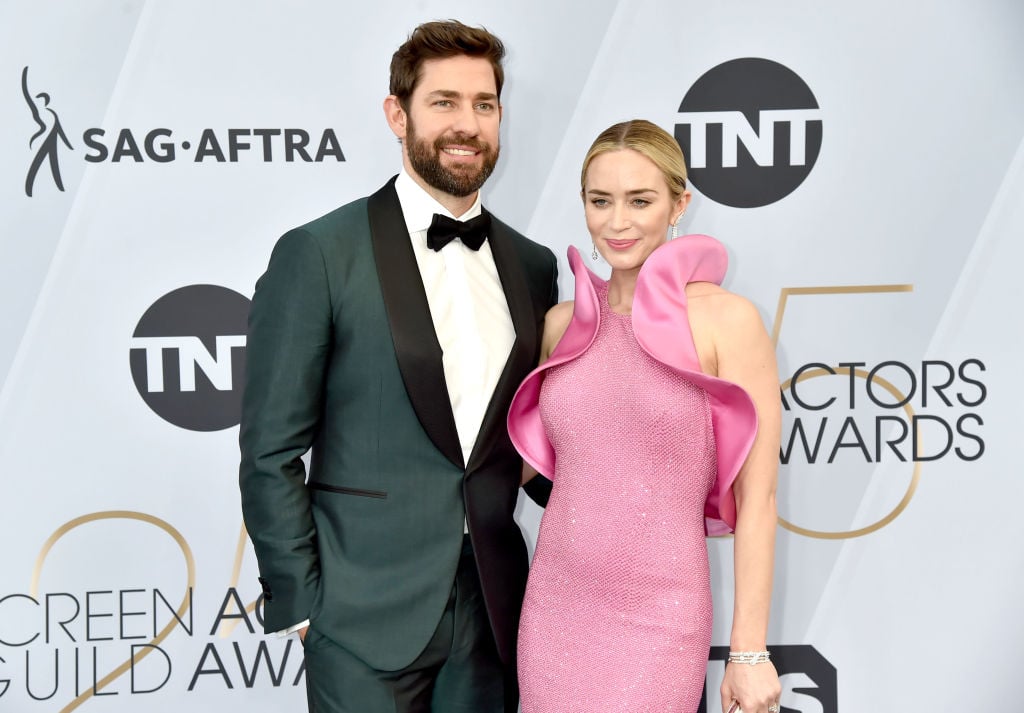 John Krasinski and Emily Blunt smiling in front of a white repeating background