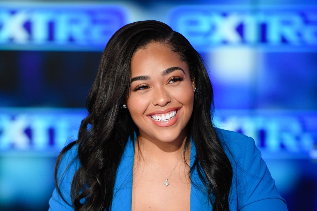Jordyn Woods smiling, turned away from the camera, in front of a blue background