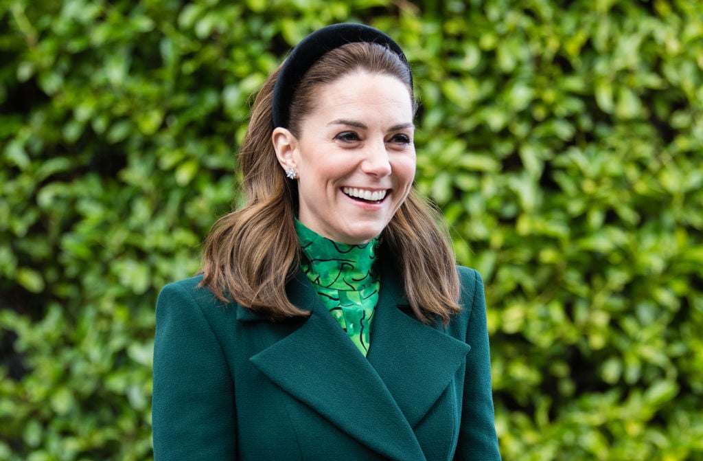 Kate Middleton smiling and looking off camera, wearing green in front of a green background