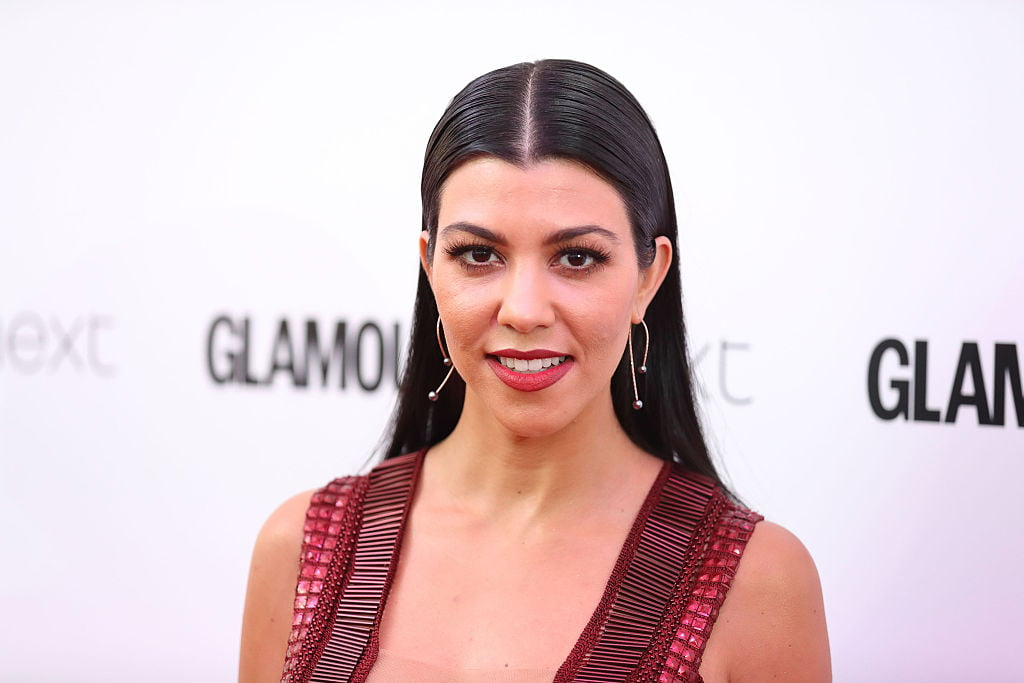 Kourtney Kardashian wearing red, smiling at the camera in front of a white background with repeating logos