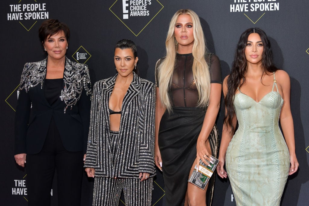 Kris Jenner, Kourtney Kardashian, Khloé Kardashian and Kim Kardashian West smiling in front of a black background with repeating logo